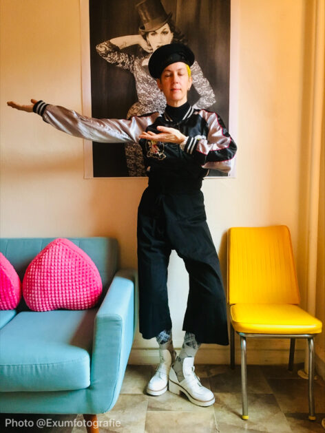 A full body photo of Claire who stands in a room with colorful furniture; they are wearing a black beret and close their eyes while their palms face upward and one arm is extended to the side. Photo by Exumfotographie.