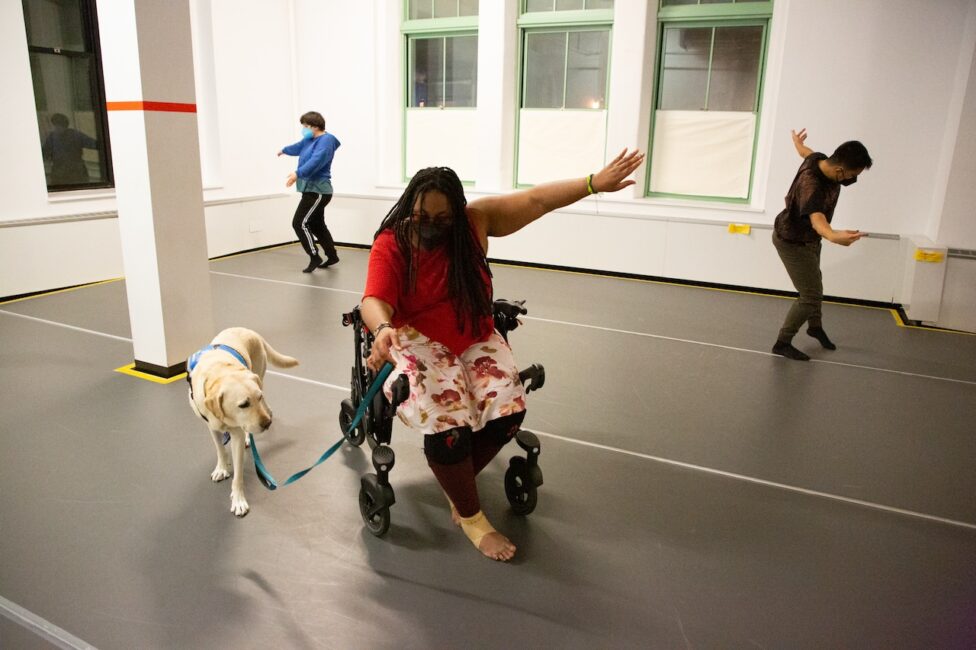 Photo from Elisabeth Motley and Kayla Hamilton’s AMP Residency showing. Three dancers move around the space. In the foreground a dancer moves on a wheelchair with their arm extended to the side accompanied by a service dog. Photo by Whitney Browne.