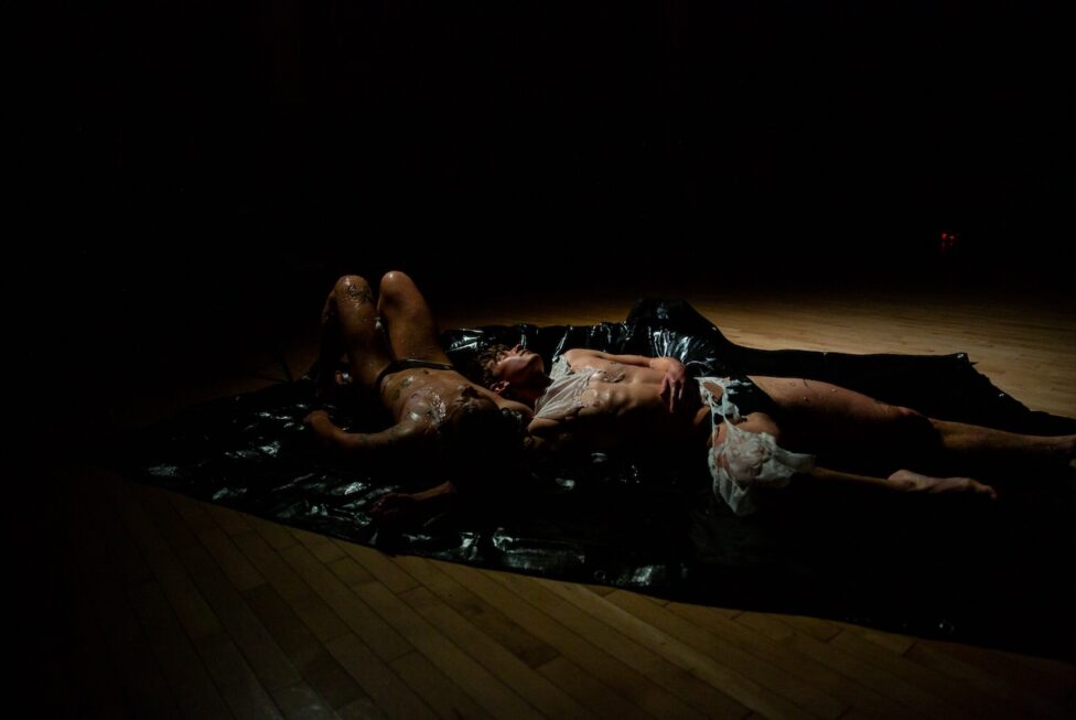 Photo by Rachel Keane, Dancers felix bryan and cove barton, MR@Judson: felix bryan and cove barton lay sprawled out with their heads near each others shoulders on a black tarp. Their white costumes are wet and dissolving into nothing.