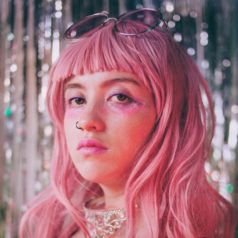 ID: The image features an artist's portrait with a retro and androgynous aesthetic, wearing a pink wig, glittery makeup, and a velvet outfit in shades of pink. The shimmering, blurred background, along with accessories like glasses and an ornate necklace, enhance a dreamy and nostalgic atmosphere. Photo taken by Luisa Ramírez.