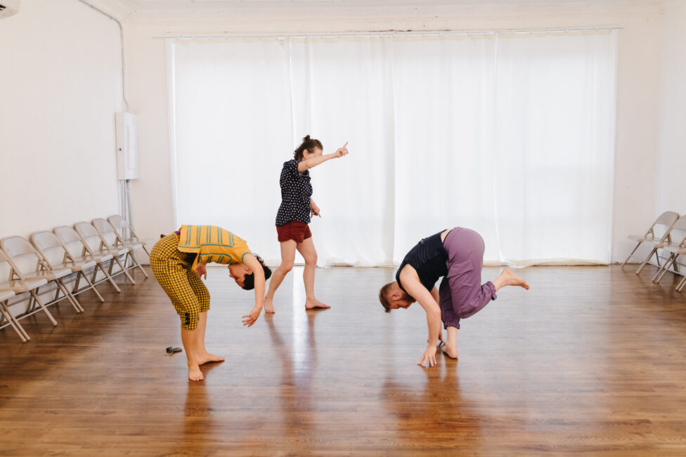Three dancers move upon an shiny wooden floor. Two of them curl over and into themselves, and one stands with right arm outstretched, index finger pointing up. Photo credit to Lory Lyon. 