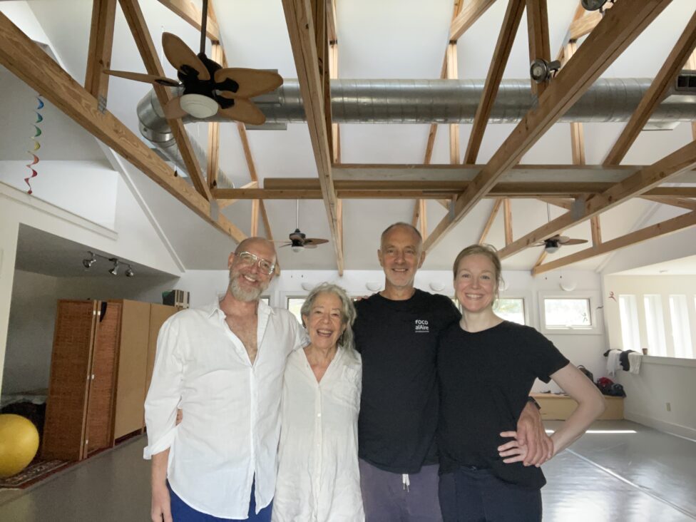 Four people pose in a dance studio, standing close to one another and smiling towards the camera. Photo credit: Christopher Roman.