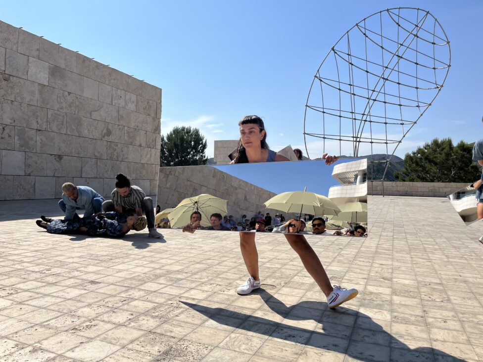 Beneath a sunny blue sky, devika holds a rectangular mirror up to reflect an audience of viewers. Photo credit to Carolyn Pennybacker Riggs. Joan Jonas,