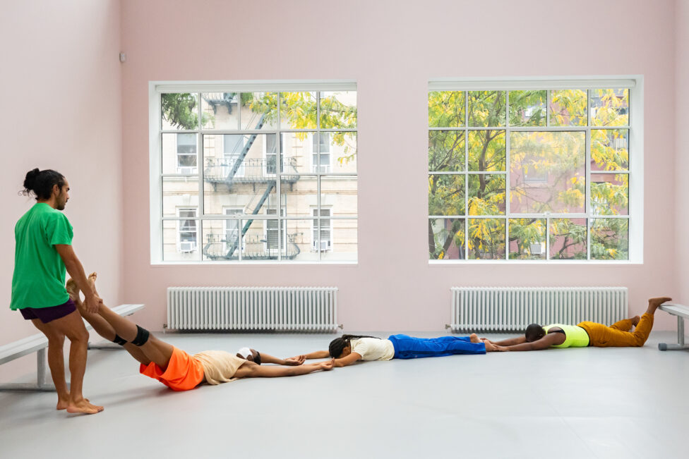 A room with pale pink walls and large windows frames four brightly costumed dancers. Three of the dancers lay horizontally across the diagonal of the floor, holding one another's limbs, while one dancer stands and holds the ankles of the last person in the chain. Photo credit to Helena Goñi. Ligia Lewis' study now steady at CARA, NYC.