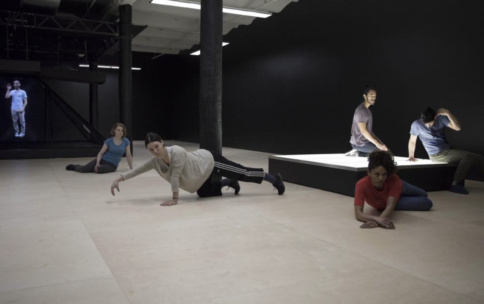 Six dancers are scattered throughout the space in various positions. The room consists of a long black wall and white floor, and two of the dancers rest upon a rectangular structure that emits a soft white light. Photo Credit to Julieta Cervantes. Alexandra Pirici's Co-Natural at The New Museum NYC.