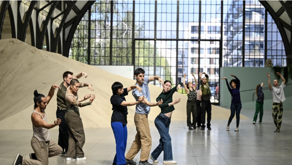 In a large space with floor to ceiling windows, many figures stand in small groupings. The figures create differing, rounded shapes with their arms, their eyes intently fixed upon their hands. Photo credit to Reto Klar. Alexandra Pirici's Attune at Hamburger Bahnhof, Berlin.