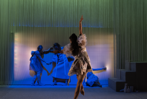 A dancer (devika) extends their right arm to the sky while perched on the ball of their right foot. Below them is a square of a reflective surface, and they face a soft green, lavender, and yellow backdrop. Photo credit to Kameron Herndon. Big Dance Theater, 