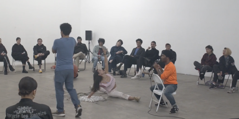Wearing a pale purple shirt and white pants, devika poses on the ground in a split, their left arm reaching upwards. Along the perimeter of a large white wall, people sit in chairs and watch. Photo credit to Ajani Brannum. Ajani Brannum,