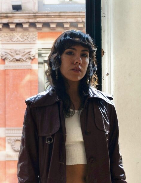 Yara: A light brown skinned Latina with long wavy brown hair looks directly at the viewer. She is in a studio with a window revealing a NYC building by day and she is wearing a burgundy trench coat and a white tank top. Photo by Blackwall (Yanasasha)
