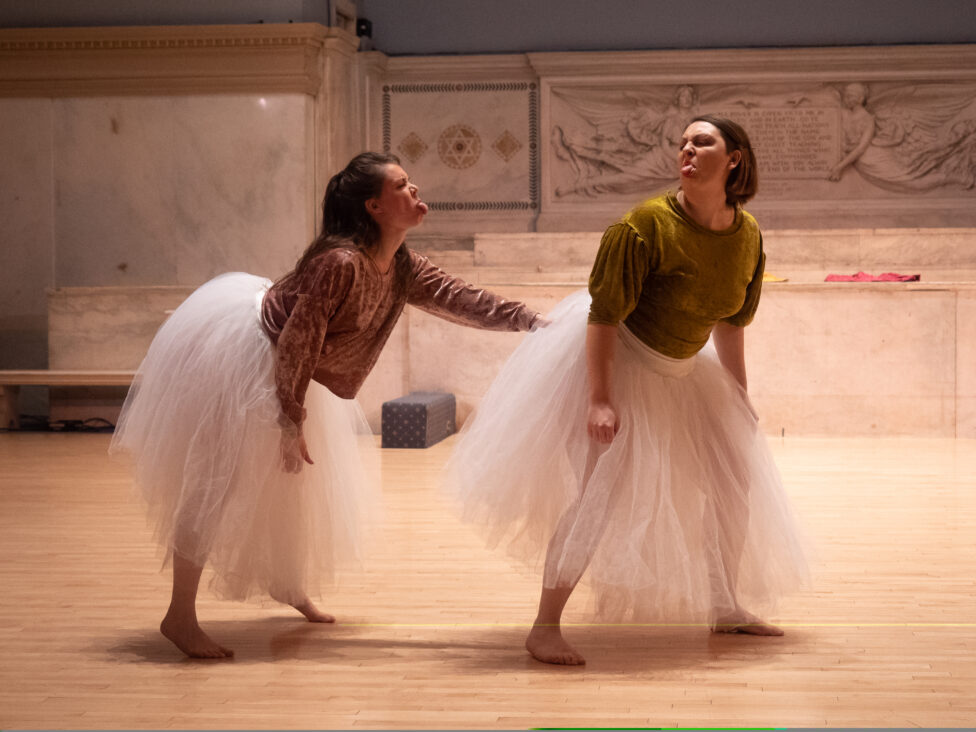 Photo from Paige Phillips’ performance for Movement Research at the Judson Church: Two dancers wear velvet tops and long light pink tulle skirts; they look towards each other sticking their tongues out. Photo by Iki Nakagawa.