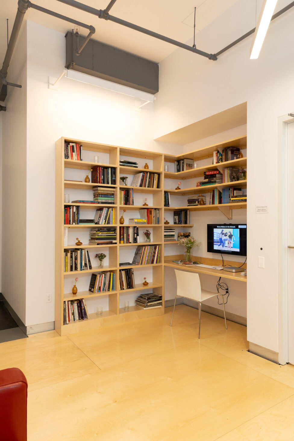 Small and cozy library space with a computer on a desk and a white chair within a corner space. There are rectangular wooden shelves with different color books on them. Daniela Garza Maldonado.