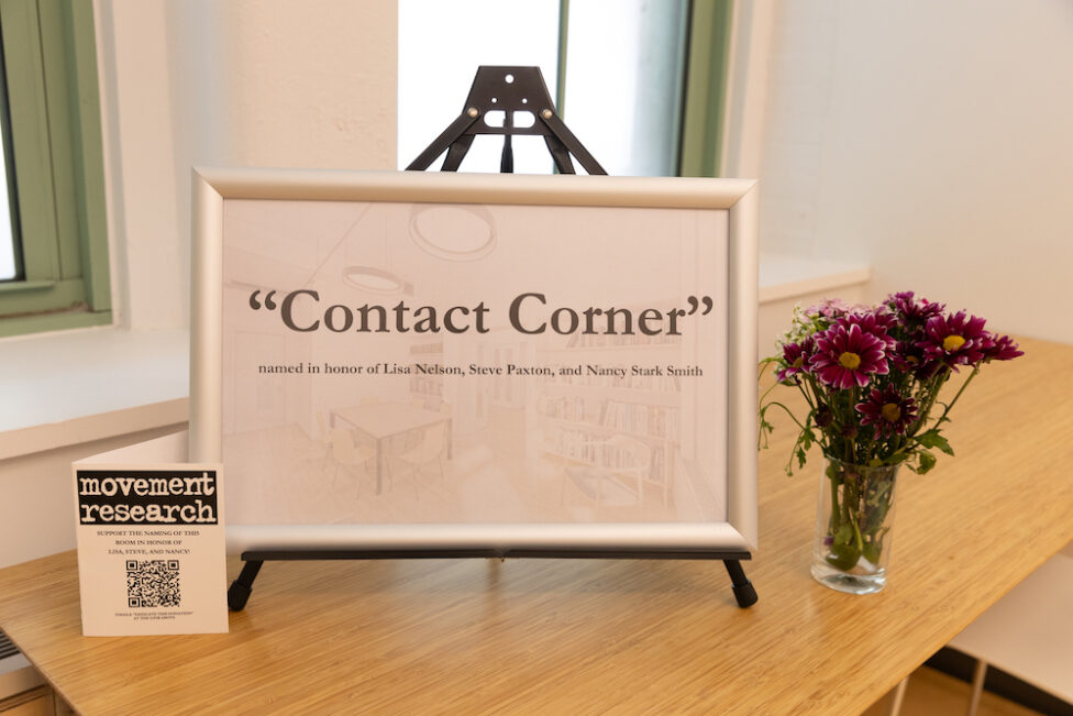 White sign with white frame on top of wooden counter that reads “Contact Corner”, below in smaller text it reads “Named in honor of Lisa Nelson, Steve Paxton, and Nancy Stark Smith”. On the left corner, a QR code with Movement Research classes and information.