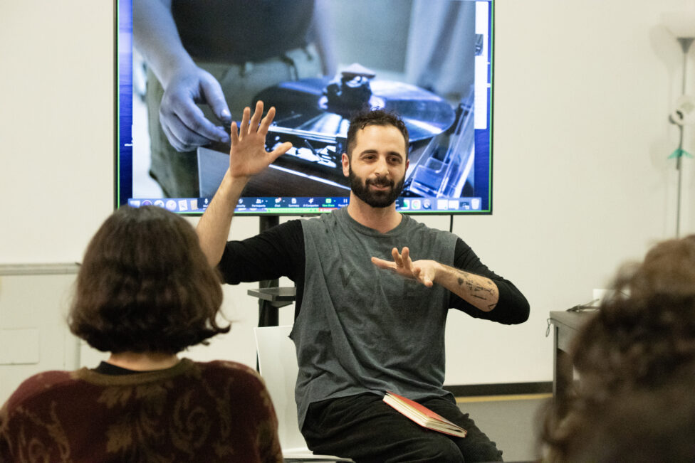 GPS Chat with Charlie Prince, “Solidarity, displacement, and inverted process in contemporary practice” A photo of Charlie sitting in front of a TV gesturing to the participants. Photo by Abigail Montes.