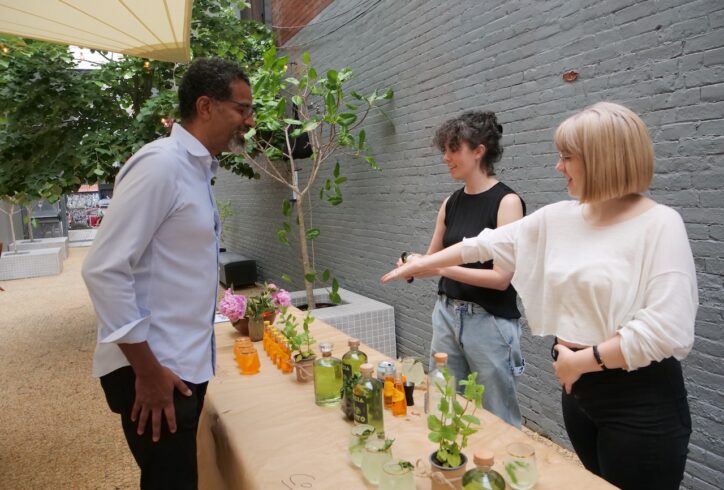 MR Staff, Leah and Elle stand behind a table in the 122CC courtyard smiling at a guest, gesturing at the drinks presented amongst flowers.