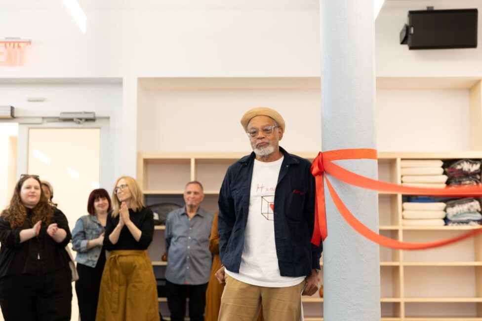 A man with light brown skin dressed in a beige flat cap, black jacket, white t-shirt and tan khaki pants leans his left side against a white pillar in a dance studio. There is an orange ribbon around the pillar that he is leaning on. There are shelves in the background with yoga blankets and another man of color standing in the frame but not In focus. Photo by Daniela Garza Maldonado.