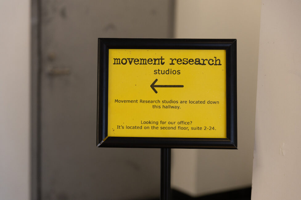 Yellow Sign with black frame that reads “Movement Research studios” with an arrow below pointing to the left. Below the arrow it reads “Movement Research studios are located down this hallway. Looking for our office? It’s located on the second floor, suite 2-24. Photo by Daniela Garza Maldonado.