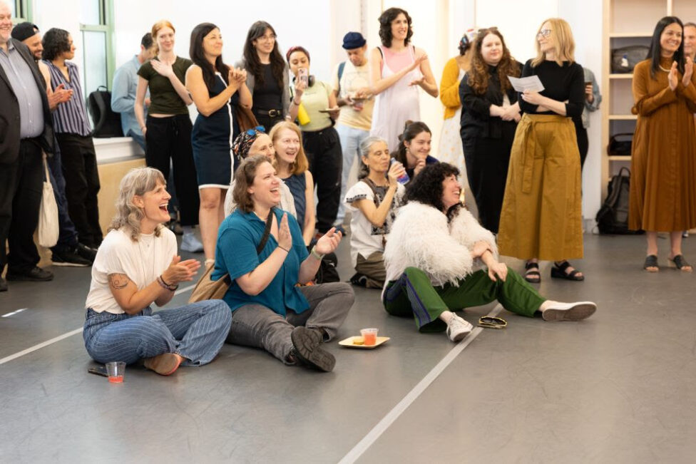 In a dance studio with a grey marley floor 23 people (mostly white, some people of color) are gathered. 7 of them are sitting, 16 of them are standing or leaning against the wall. They are applauding and have cheerful expressions. Photo by Daniela Garza Maldonado.