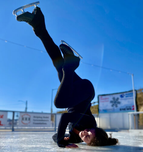 On an outdoor ice rink with a clear blue sky above, a white, female bodied person in their mid 20's does a shoulder stand on the ice on their left shoulder. Their right hand is supporting them and their legs are in the air, their right leg is straight while their left leg is bent at the knee. They are dressed in all black with black ice skates.