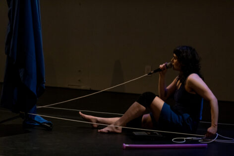 Allie, a white woman with dark hair, sits on the ground in a black box theater speaking into a microphone with a white mic cable.   Photo by Michelle Smith Lewis. 