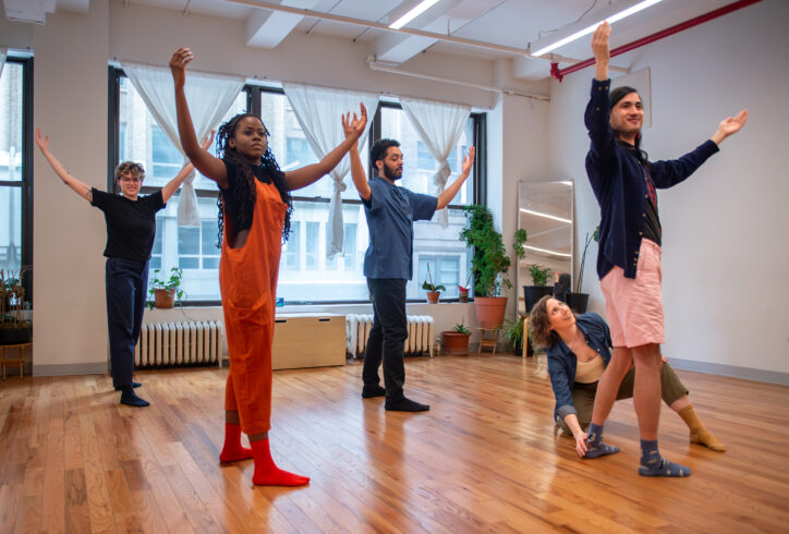 A group of dancers in a studio stand with one leg forward and arms raising upwards. Allie crouches on the ground adjusting a dancer's foot. Photo courtesy of the artist.
