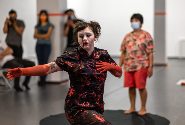 OP, 2022 at 122CC; An artist with white paint on their face and red streaking down one eye wears a black Qipao with red flowers; their hands and forearms are covered in red paint as well, while they place one hand to their chest and the other stretched out; in the background 4 masked audience members observe. Photo by Rachel Keane.