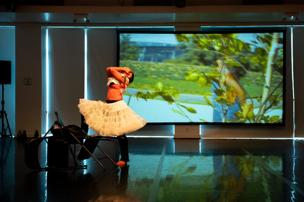 Symara stands with both hands pressed to her face, head tilted up. She wears a billowing white petticoat. Behind her is a projection image of green leaves and a natural landscape. Image by Maria Baranova.