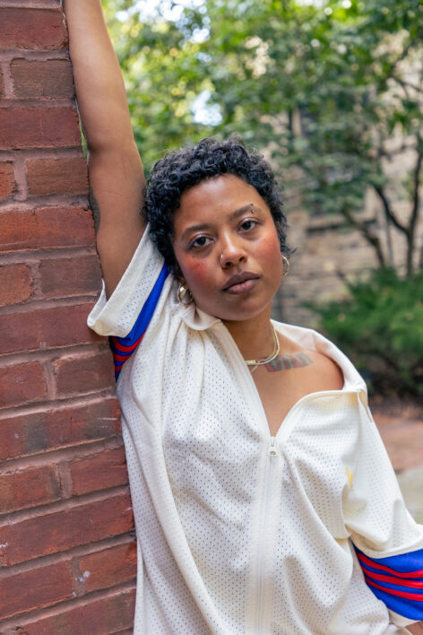 A brown femme with short curly black hair is in a blue and white puffer and sparkly baggy jeans with her arms opened to the side with one foot propped in a demi releve and the other foot firmly planted on the floor. She is saturated in a deep red light that envelops the whole space.Photo by Elyse Mertz courtesy of Center for Performance Research.