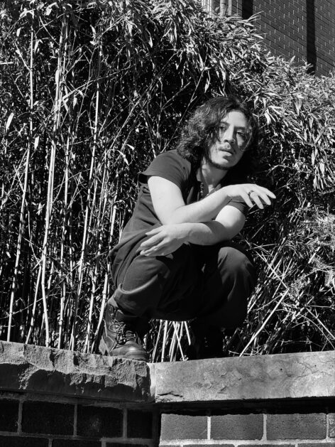A black and white photo of Edu Tair crouching on a brick edge with bamboo branches cast as a background. His/Their hair is down and wearing black clothes with a black ring. Photo/Image by Edu Tair Hernandez.