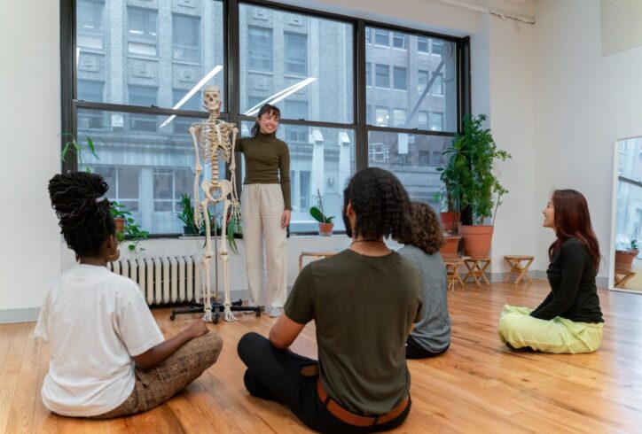Holly is smiling and touching the spine a life-sized human skeleton as she faces a class full of students who are seated on the floor. Photo by Bryan Bisordi.