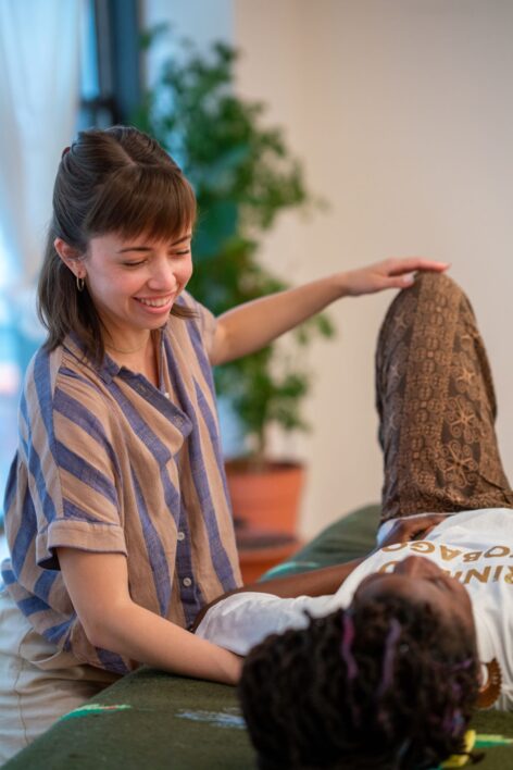 Holly’s Alexander student lays on a green massage table. Holly stands next to the table and is smiling as she lightly touches her student’s knee and shoulder with her hands. Holly’s Alexander student lays on a green massage table. Holly stands next to the table and is smiling as she lightly touches her student’s knee and shoulder with her hands. Photo by Bryan Bisordi