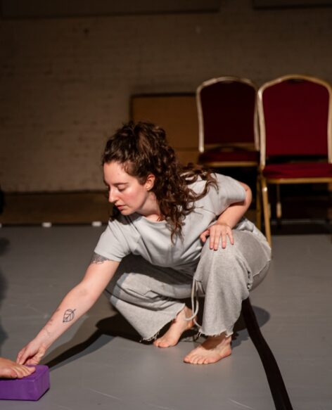 Sofia, a white brunette dressed in loose grey athletic gear, squats low to the ground. She reaches one arm toward a purple yoga block that is at the edge of the frame. The floor is grey and empty maroon chairs sit behind her. Photo by Rachel Keane.