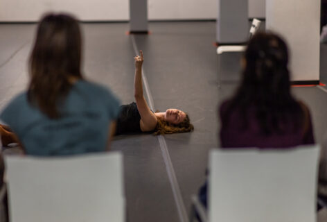 A photo of an Open Performance artist reaching their arm up and laying on the ground. In the foreground the audience turns its back tot he camera viewing the performance.