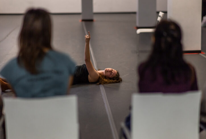 A photo of an Open Performance artist reaching their arm up and laying on the ground. In the foreground the audience turns its back tot he camera viewing the performance.
