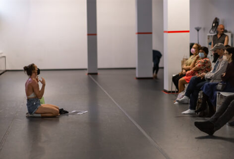 Photo of an open performance artist sitting and gesturing with their hands. The audience watches attentively. Photo by Rachel Keane