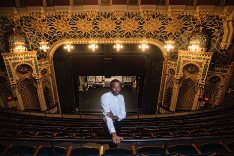 Antuan Byers stands confidently in the balcony of New York City Center theater, dressed in a crisp white shirt and black slacks. The rich architectural details, including golden geometric patterns, arched motifs, and glowing chandeliers, frame the space in grandeur. Behind him, the empty stage and rows of seats evoke a sense of both legacy and possibility. His poised presence, centered in a space of performance and power, reflects his dual role as an artist and organizer. The image captures a moment of reflection and vision, symbolizing his commitment to transforming the dance world from the inside out. Photo by Eric Politzer