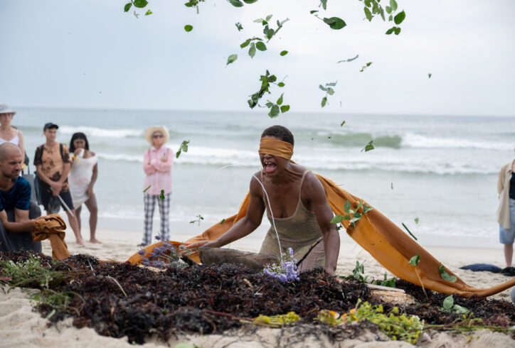 black artist kneeling in the sand on a beach with an elongated golden blindfold surrounded by a small group of people. Ocean in the background. Artist siittting in front of a mound of seaweed and plant parts captured in a gesture of throwing, leaves suspended overhead. Dynamic colors of ocean blue, green leaves, brown sand, the artist brown skin and the silver shimmer of the sun on the water in the background. Nir Arieli.