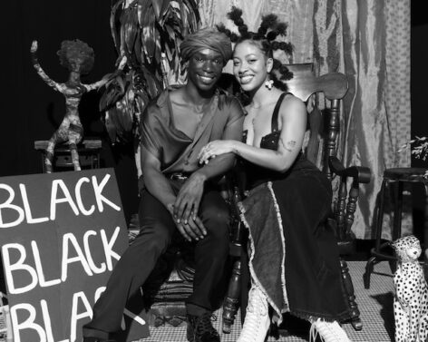 A black woman and a black man sitting side smiling at the camera with a sign to their left that says “Black Black Black” photo by Geoffrey Baptiste