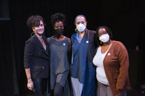 Against a black background we see four people standing looking at the camera, arms around each other’s backs. From left to right we see Paloma McGregor, butterscotch skinned, wearing a black and white pinstriped suit with hoop earrings and curly black hair in a ponytail. Followed by Marguerite Hemmings deep cocoa skinned wearing a black face mask, grey checkered sleeveless top with black pants, hair is in a messy bun. Followed by Joya Powell butterscotch skinned wearing a white face mask, glasses, long flowy multi-patterned jumpsuit. Followed by Kayla Hamilton, caramel skinned, wearing a white mask, glasses, brown knitted cardigan over a white shirt and mud colored linen pants. Photo by Whitney Browne.
