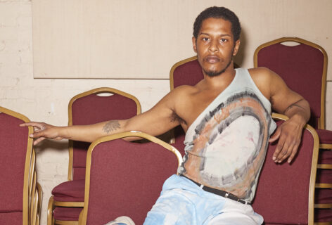 Malcolm sits on a stack of red chairs, he looks direct to the camera leaning slightly right. Photo courtesy of the artist.