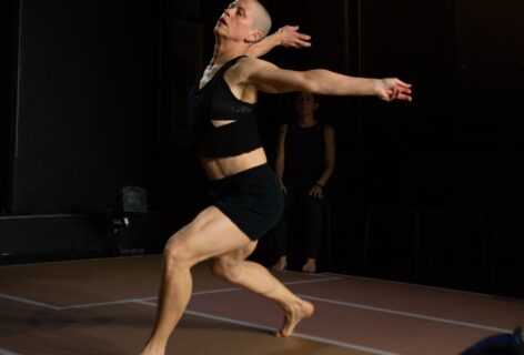 a white woman with a shaved head, wearing blue sweatpants and a black and gray shirt, moves through phrase material, leaning into right hip, left leg lifted and bent, elbows bent while the right swings forward and the left arm swings back. Photo by Rosa Wolff.
