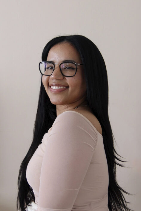 Leomary Rodriguez Headshot by Leomary. A photo of Leomary looking over her shoulder at the camera smiling, she is wearing a sheer top and black framed glasses.