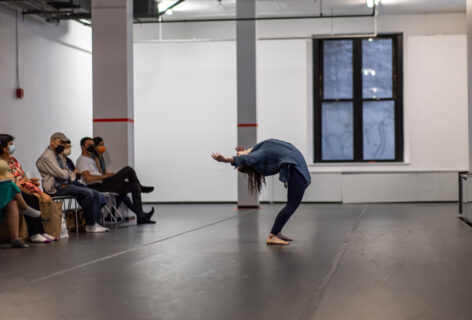 A photo of an Open Performance artist standing in a deep back bend reaching towards the onlooking audience. Photo by Rachel Keane