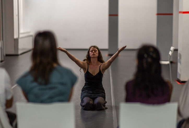 An Open Performance artist sits with their arms stretched out and upward. In the foreground the audience turns its back tot he camera viewing the performance. Photo by Rachel Keane.
