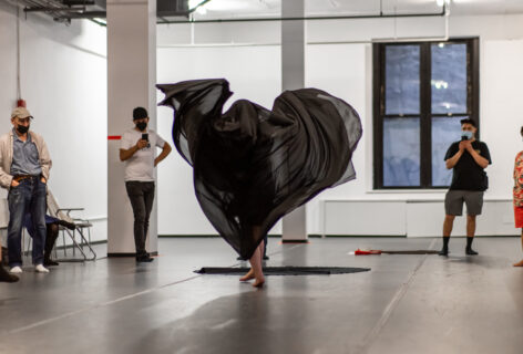 A photo of an Open performance artist. The artist stands in the middle of the apace. A sheer black fabric which is draper over their body billows out all around them. Photo by Rachel Keane.