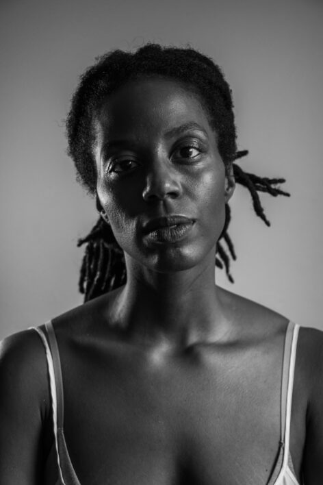 A black and white photo of a deep cocoa skinned person with locs pulled back in a messy bun looking at the camera with a serious and soft expression. Photo by Dante Napoli.