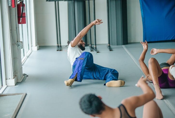 A black man lays on the ground mid movement with a smile. He is surrounded by two other dances. Photo by Nicole Miller
