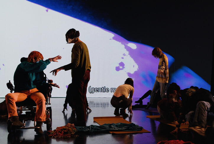 A number of masked dancers, one wheelchair user, some people, standing, some lying down, are moving together in front of a projection of water in pink and white. Photo by Petra Kuppers.
