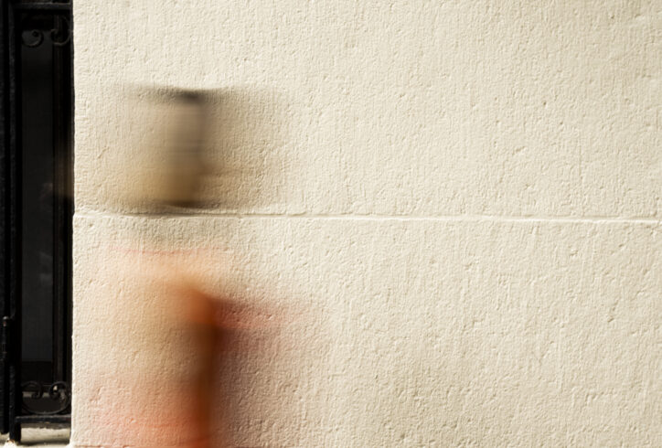 The photo shows a blurred figure in motion walking past a textured, beige wall. The person is wearing a red outfit, with their head and body smeared across the image as a result of a slow shutter speed. On the left side of the image, there is a black window with vertical iron bars, contrasting against the smooth wall and the movement of the figure. Photo By Rojin Shafiei.