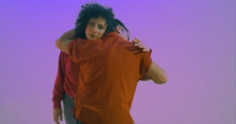 Two dancers stand, physically wrapped around each other, one with their back to the camera and the other facing out. Video Still by Chico Eastridge.