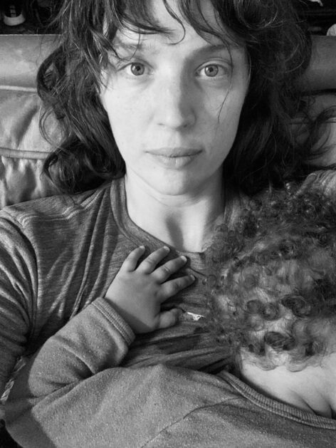 black and white close up of white woman with shaggy brown hair gazing directly at camera, with curly haired infant resting on her chest.